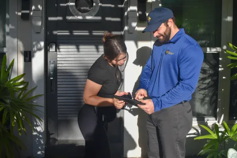 Woman and pest control man reviewing a tablet outside a residential front door on a sunny day.