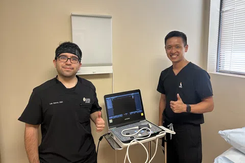 Two male medical professionals in black scrubs giving thumbs up next to an ultrasound machine in a clinic room.