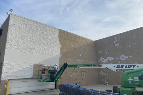 Workers repainting a large beige building wall white using a JLG lift and spray equipment under a blue sky.