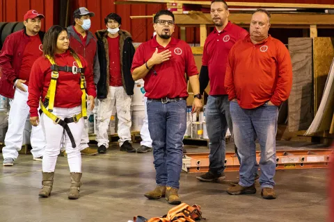 Group of workers in red shirts in a warehouse, one wearing a safety harness and others listening attentively.