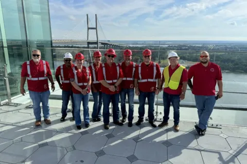 Group of construction workers wearing safety gear posing on rooftop with bridge and river in background.