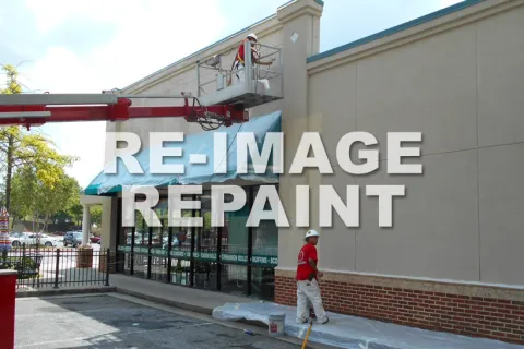 Construction workers use lift equipment to paint the exterior of a commercial building near a parking lot.