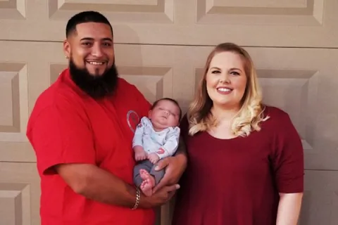 Smiling couple stands in front of garage door holding their newborn baby in casual outfits on a sunny day.