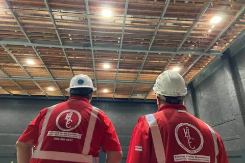 Two construction workers in red safety vests and helmets inspecting a ceiling in an industrial building.