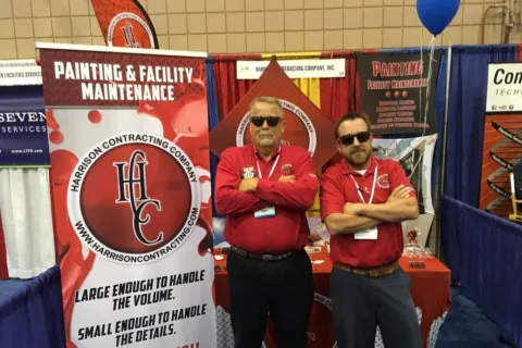 Two men in red shirts and sunglasses stand with arms crossed at Harrison Contracting Company booth with painting and maintenance banners.