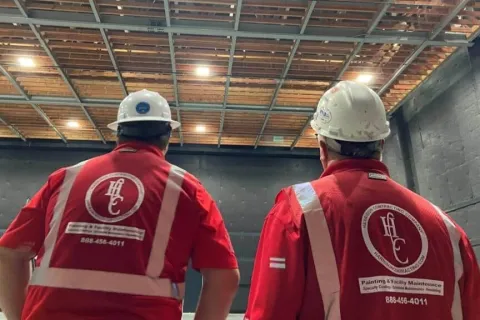 Two workers in white helmets and red safety vests inside a large industrial space with exposed ceiling beams.