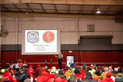 Speaker presenting 2022 Safety Day to a large indoor audience wearing red and yellow safety gear in a warehouse.