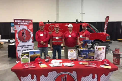 Four men wearing red shirts and hard hats stand behind a red table with promotional items and a van at a trade show booth.