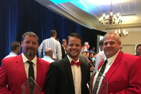 Three men at award ceremony, two wearing red blazers holding trophies, one in black suit with red bowtie and sneakers.