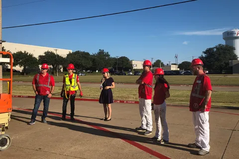 Group of workers and a supervisor wearing red helmets and safety gear standing outdoors near a scissor lift on a sunny day