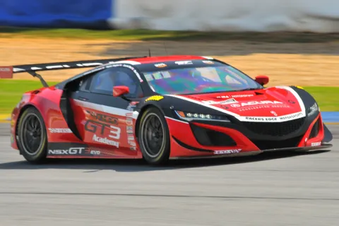 Red and black GT3 race car speeding on a racetrack with visible sponsor logos and aerodynamic design.