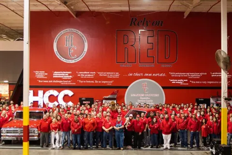 Large group of employees in red shirts posing in front of red wall with HCC industrial contracting slogans and logos