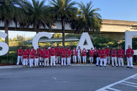 Group of people in red vests and white pants standing in front of large SCAD letters with palm trees and overpass behind.