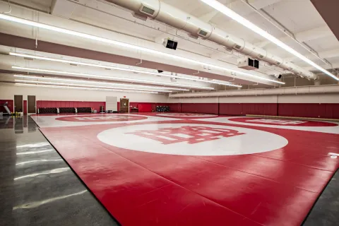 Indoor wrestling gym with red mats featuring large white circles and a logo, bright overhead lighting.