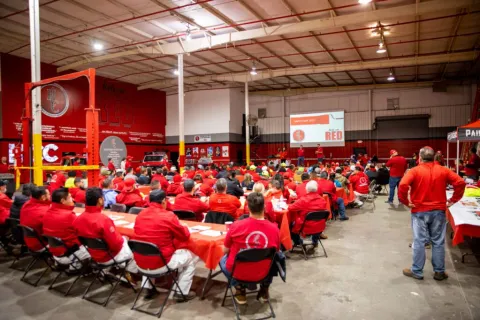 Large group of workers in red uniforms attending safety training session in a spacious industrial warehouse.
