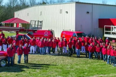 Group of workers in red shirts standing outside a warehouse with red service trucks and equipment on a grassy area.