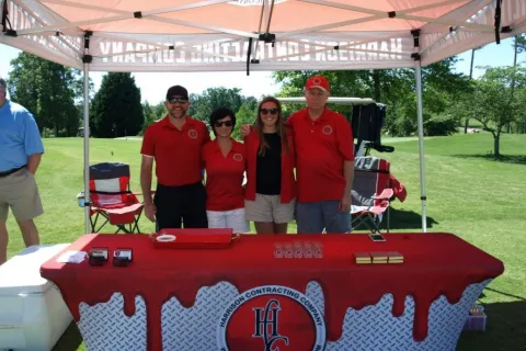 Four people in red shirts stand behind a red table with Harrison Contracting Company logo at outdoor event under a canopy.