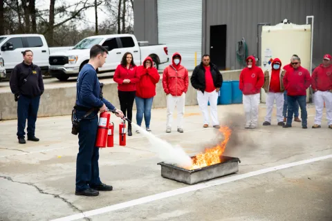 Person using fire extinguisher to put out a controlled fire during outdoor safety training with group observing.