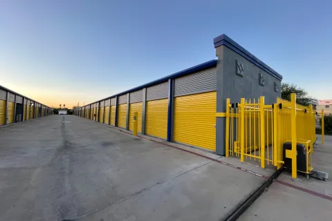 Wide view of a storage facility with yellow roll-up doors and gated entrance under a clear sky at sunset.