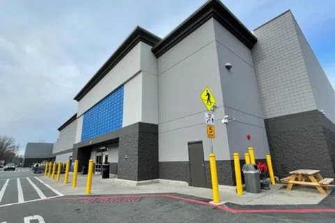 Modern commercial building with gray and blue exterior, yellow safety poles, and pedestrian crossing signs in parking lot.