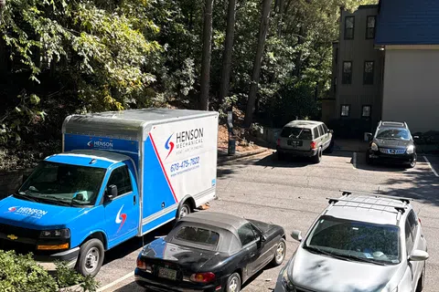 Blue and white Henson Mechanical service truck and three cars parked in a wooded apartment lot on a sunny day.