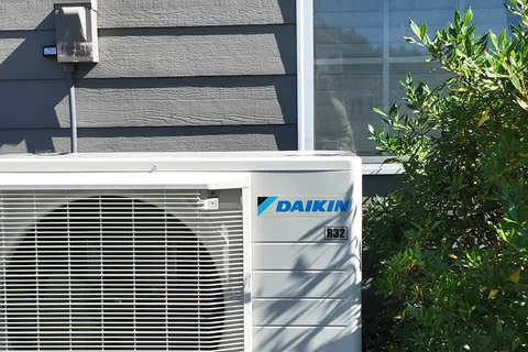Daikin R32 air conditioning unit installed outside a gray house near a window and green shrubbery.