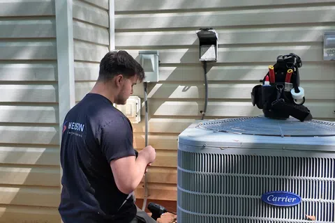 Technician kneeling beside a Carrier air conditioning unit outside a house during repair work.