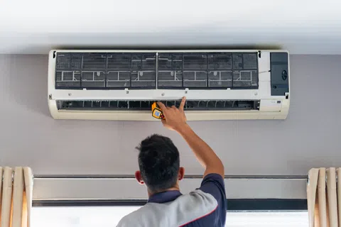 Technician inspecting and repairing a wall-mounted air conditioner unit indoors using a handheld tool.