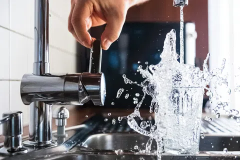 Hand turning faucet on as water splashes out of a glass in a kitchen sink, motion captured mid-splash.