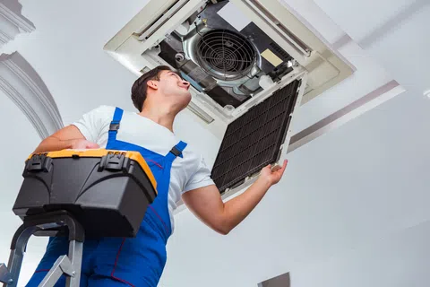 Technician in blue overalls inspecting and repairing ceiling air conditioning unit while holding a black toolbox
