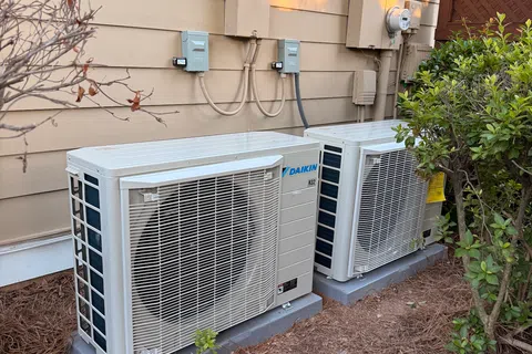 Two outdoor Daikin air conditioning units installed on a beige house exterior with nearby plants and pine straw mulch.