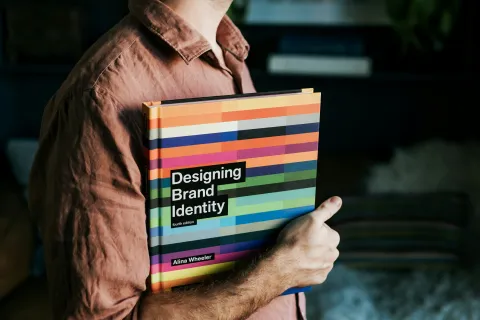 Person holding a colorful book titled Designing Brand Identity by Alina Wheeler in a cozy room.