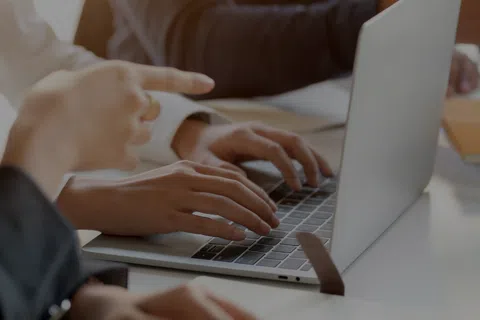 Close-up of hands typing on laptop keyboard with another person pointing at screen during meeting.
