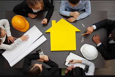 Overhead view of six architects discussing house plans around a table with a yellow house model and helmets.