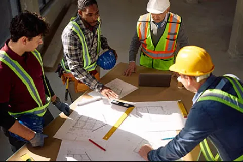 Four construction workers wearing safety gear reviewing blueprints and plans on a table at a building site.