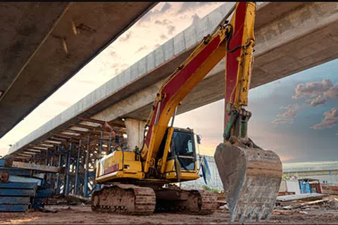 Yellow excavator with large bucket at construction site under elevated bridge during sunset.