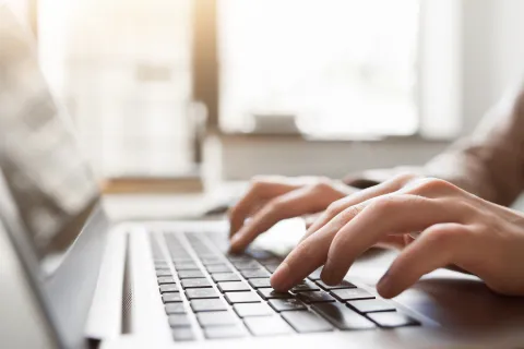Close-up of hands typing on a laptop keyboard with sunlight in the background, workspace setting.
