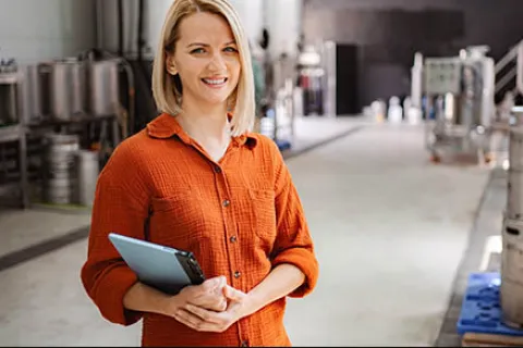 Smiling woman in orange shirt holding tablet in a brewery surrounded by metal kegs and brewing equipment.