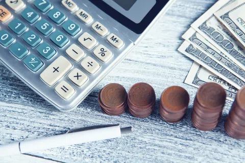 Calculator, stacks of coins, U.S. dollar bills, and a pen on a gray wooden surface representing finance and budgeting