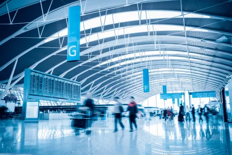 Busy modern airport terminal with travelers walking under curved ceiling beams and large flight information displays.