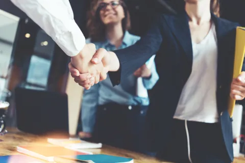 Two business professionals shaking hands in an office with a smiling colleague in the background.