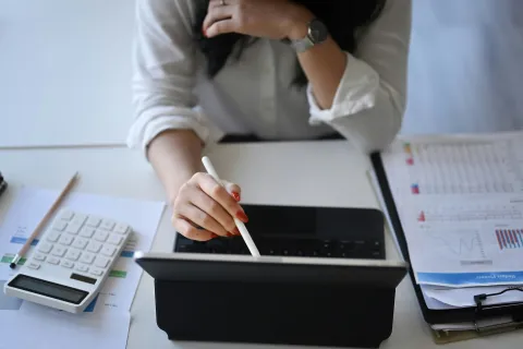 Person using a stylus on a tablet at a desk with PII documents, calculator, and notepads.