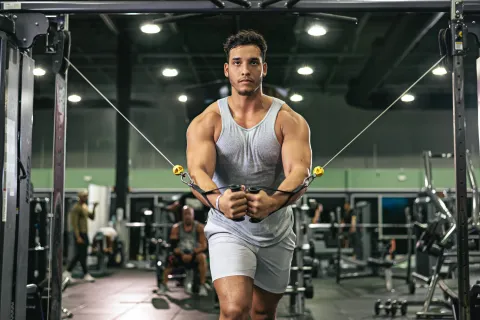 Muscular man in gray tank top and shorts performing cable chest fly exercise in gym