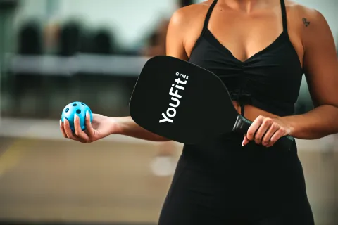 Woman in black sportswear holding a YouFit gym paddle and blue pickleball ball outdoors.