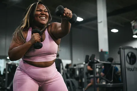 Smiling woman in pink workout clothes lifting dumbbells in a gym during strength training session.