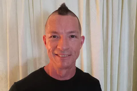 Smiling man with a mohawk haircut wearing a black YouFit Gyms t-shirt in front of white curtains.