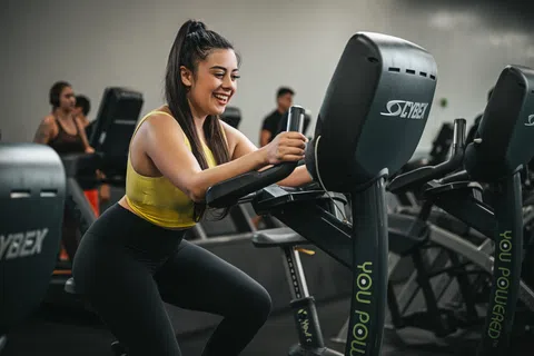 Young woman in yellow sportswear smiling while using a Cybex exercise bike in the gym.