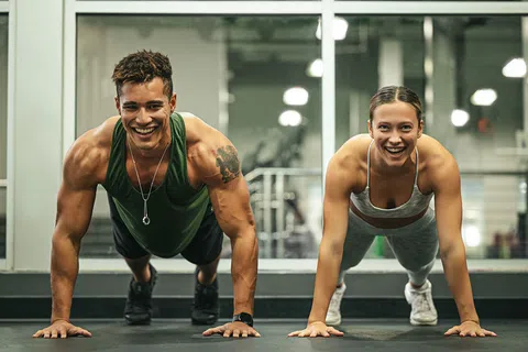 Young man and woman doing push-ups together in a gym with smiles, wearing athletic clothing.