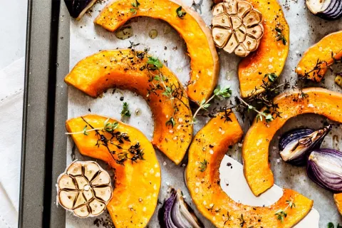 Roasted pumpkin slices with garlic, red onion, and fresh thyme on a baking tray seasoned with pepper and salt.