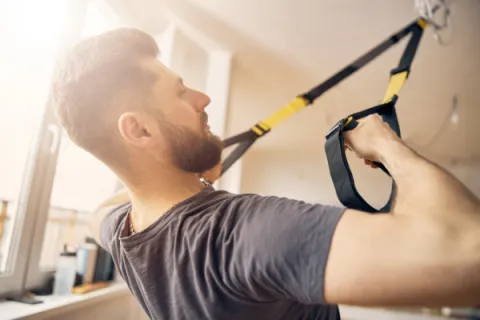 Man with beard exercising using suspension trainer straps in a bright indoor gym space.
